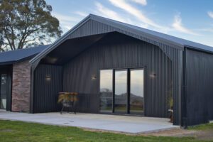 "Modern black corrugated steel building with a gabled roof, large sliding glass doors, and a concrete patio, set against a backdrop of trees and a blue sky.